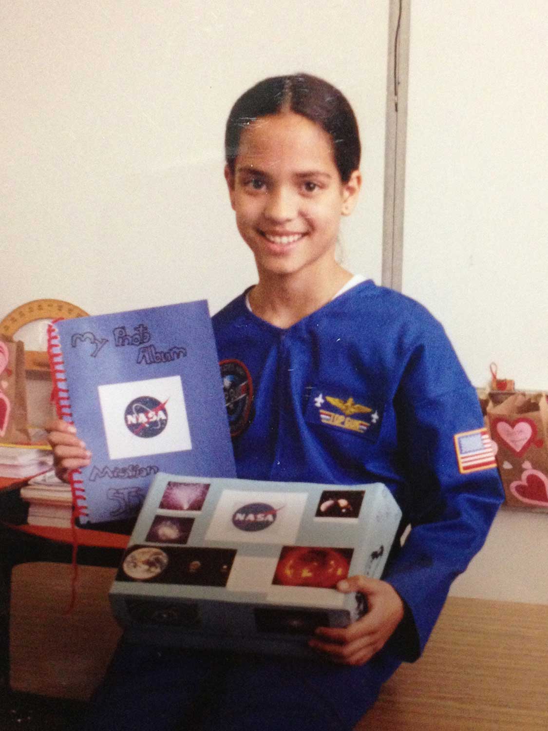 Natasha Batalha as a smiling child, wearing an astronaut flight suit and holding a scrapbook full of NASA- and space-related images.